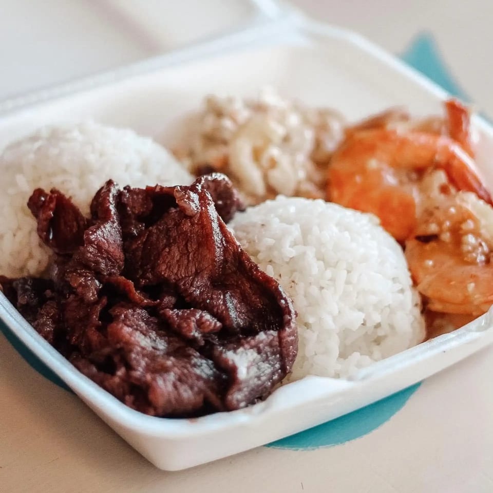 A Hawaiian plate lunch with grilled meat, white rice, macaroni salad, and shrimp in a takeout container.