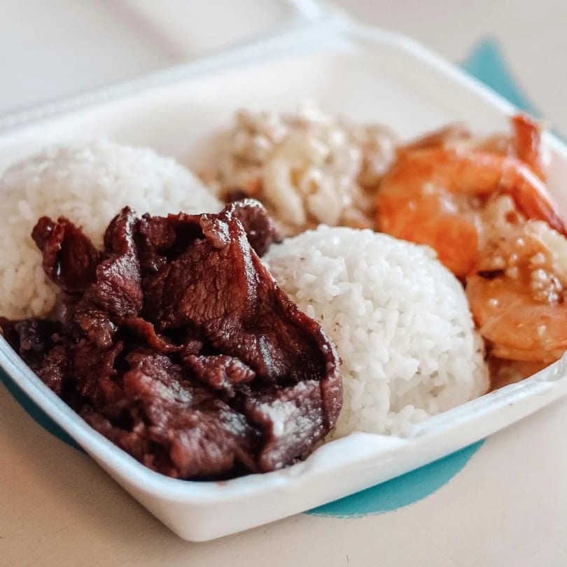 A Hawaiian plate lunch with grilled meat, white rice, macaroni salad, and shrimp in a takeout container.